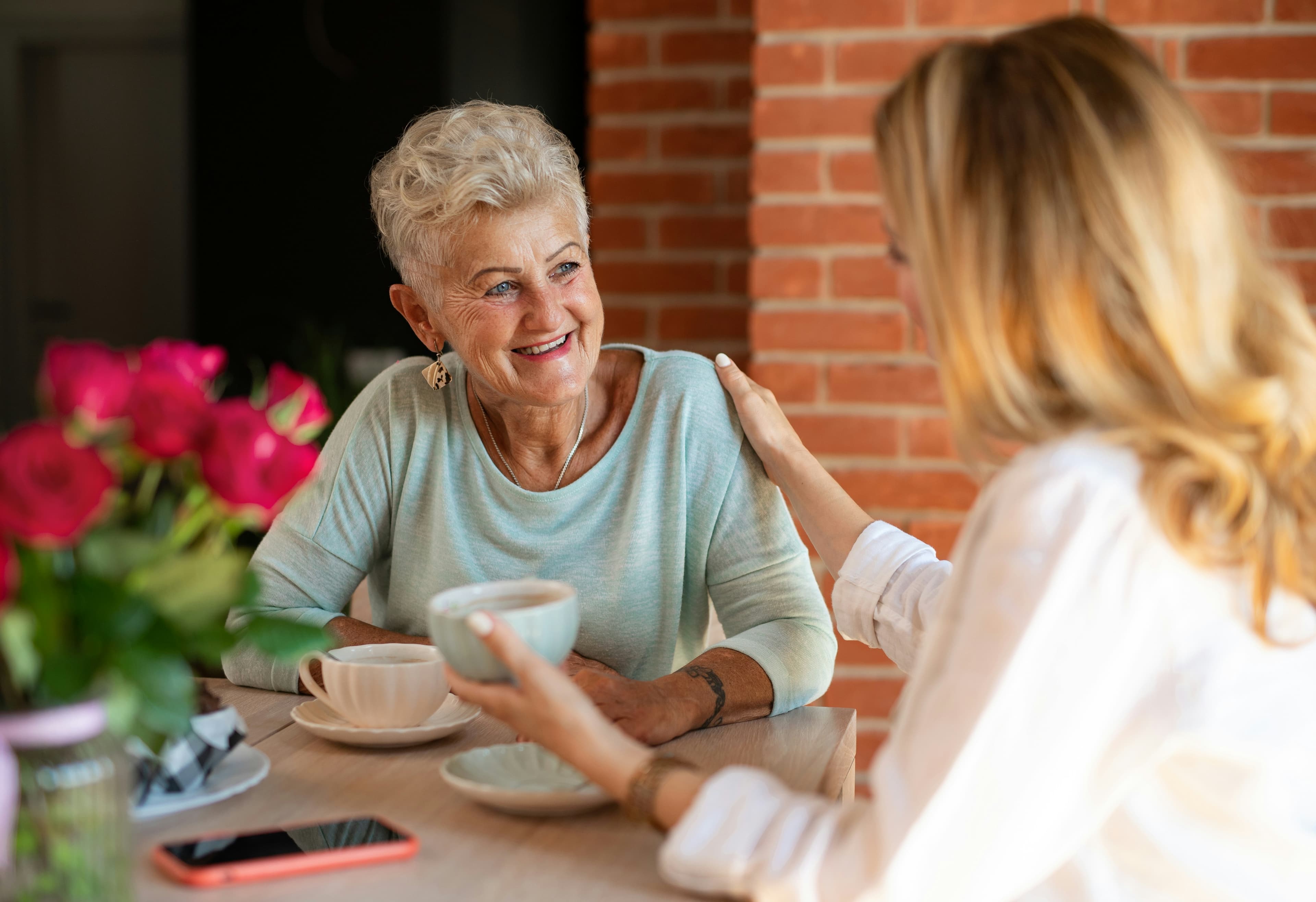 An adult daughter with her hand on her senior mother's shoulder, both smiling over coffee